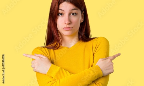 Young redhead girl with yellow sweater pointing to the laterals having doubts. Undecided person on isolated yellow background
