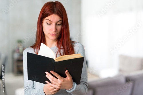 Young redhead girl holding a book and enjoying reading on unfocused background