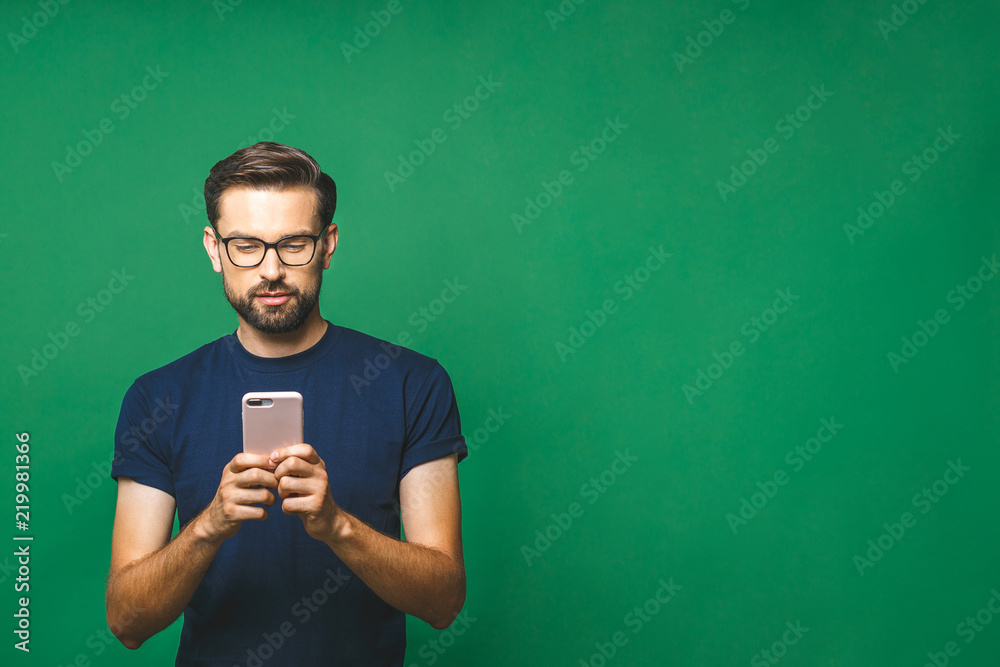 © denis_vermenko - Always in touch. Smiling young man holding smart phone and looking at it. Portrait of a happy man using mobile phone isolated over green background.