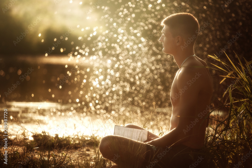a young man with a naked torso in black shorts sitting on the river bank with a book and medetiruet on the sunset