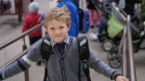 Little Caucasian curly boy in school uniform with backpack comes up on stairs.