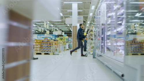 At the Supermarket: Man in a Hurry Pushes Shopping Cart full of Items, He's Walking Through Different Section of the Big Bright Mall. Following / Moving Side view Footage.