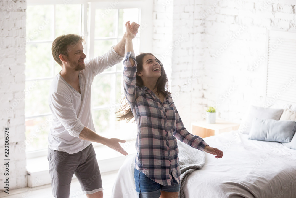 Happy funny millennial couple dancing waltz joking in bedroom holding hands and smiling, excited lovers swirl glad to move to new flat or own apartment, young couple having fun at home together Happy funny millennial couple dancing waltz joking in bedroom holding hands and smiling, excited lovers swirl glad to move to new flat or own apartment, young couple having fun at home together