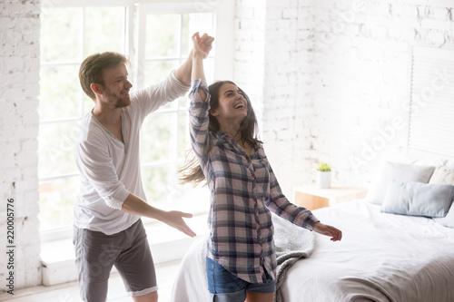 Happy funny millennial couple dancing waltz joking in bedroom holding hands and smiling, excited lovers swirl glad to move to new flat or own apartment, young couple having fun at home together