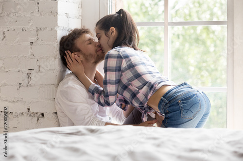 Tablou pe pânză Sensual millennial girlfriend kissing loving boyfriend sitting on windowsill at