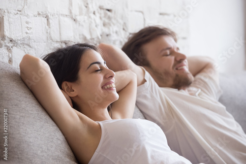 Happy millennial couple lying on cozy couch with eyes closed dreaming or imagining bright future, smiling lovers relaxing on sofa at home, enjoying rest on weekend, feeling positive relieving stress