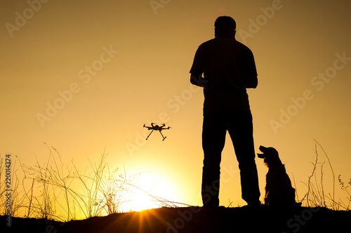 man is driving the dron at sunset the dog is looking at the drone