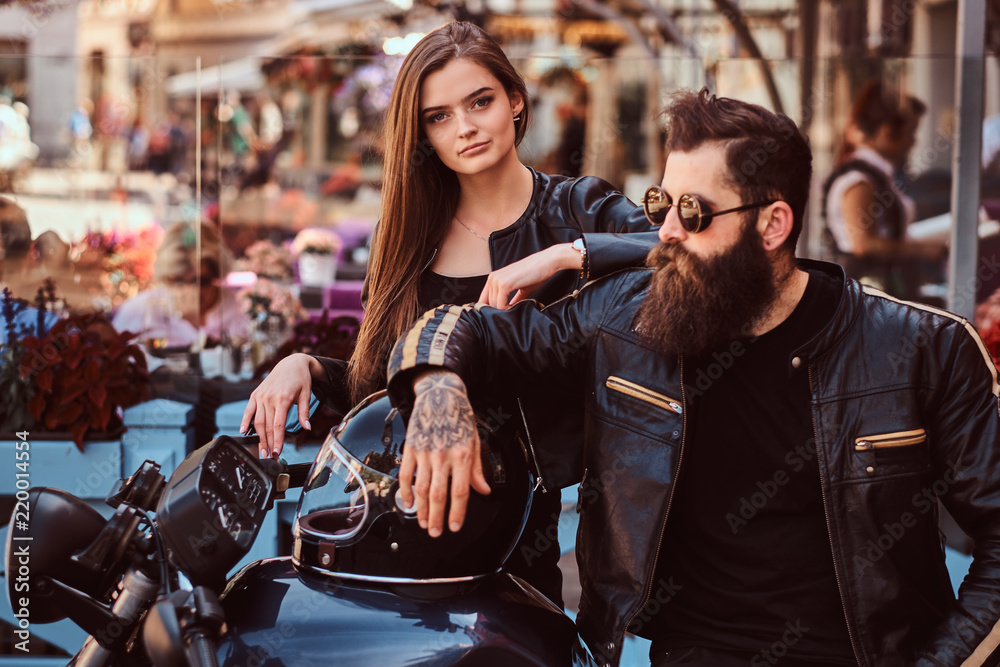 Close-up portrait of a hipster couple - bearded brutal male in sunglasses dressed in a black leather jacket and his young sensual girl standing near, posing against terrace of a cafe.