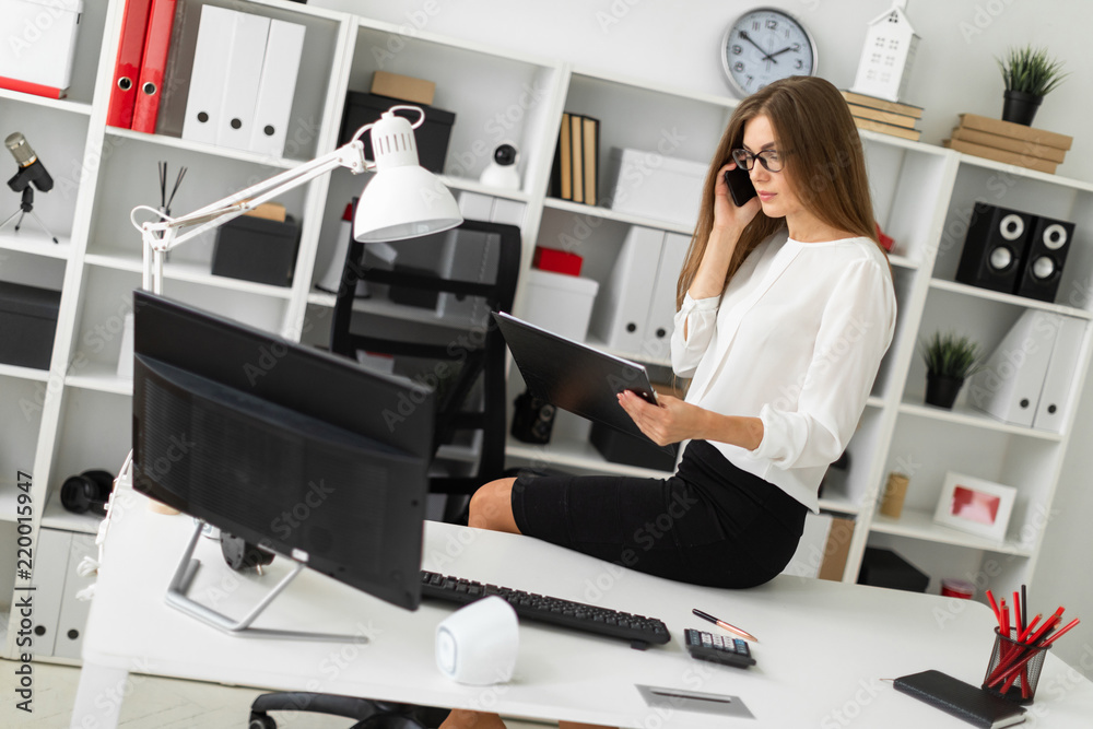The young girl sat down on the table in the office, holding a tablet with sheets in her hand and talking on the phone.