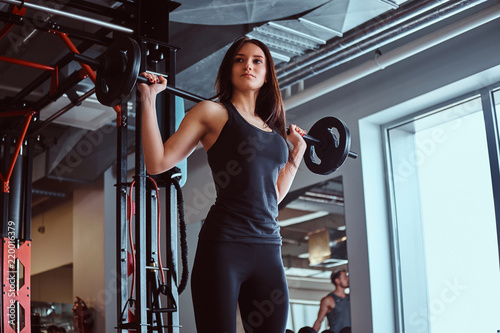 Beautiful brunette female in sportswear holds a barbell while training in a fitness club or gym.