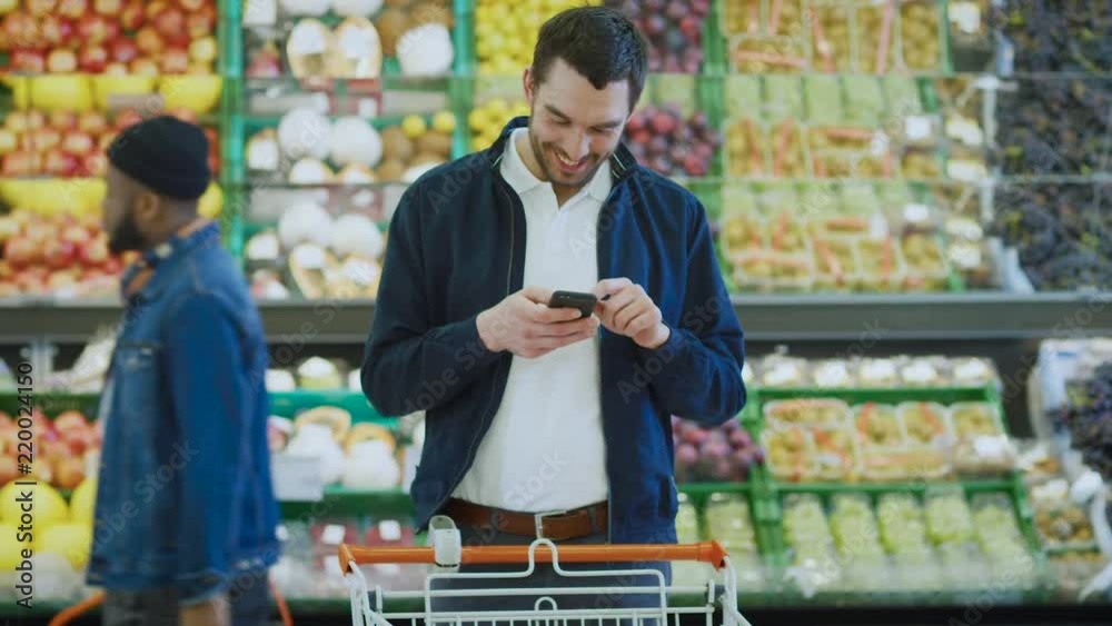 At the Supermarket: Handsome Man Uses Smartphone while Standing in the Fresh Produce Section of the Store. Man Immersed in Internet Surfing on His Mobile Phone In the Background Colorful Fruits At the Supermarket: Handsome Man Uses Smartphone while Standing in the Fresh Produce Section of the Store. Man Immersed in Internet Surfing on His Mobile Phone In the Background Colorful Fruits