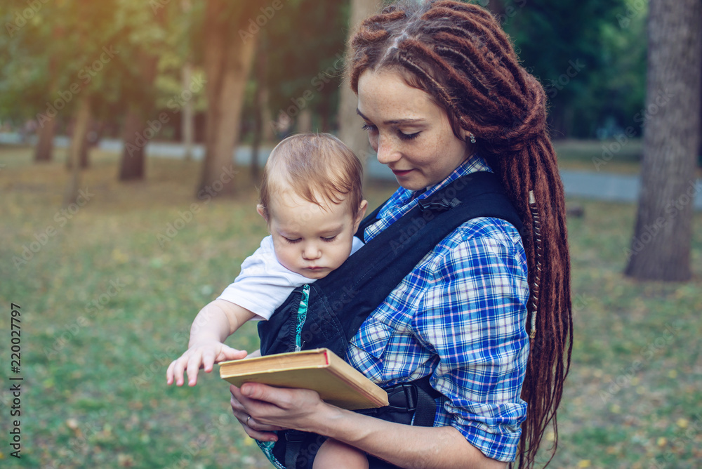 Happy mom with a baby in ergo backpack reading a fairy tale in the Park. Concept of children's education in family life Happy mom with a baby in ergo backpack reading a fairy tale in the Park. Concept of children's education in family life