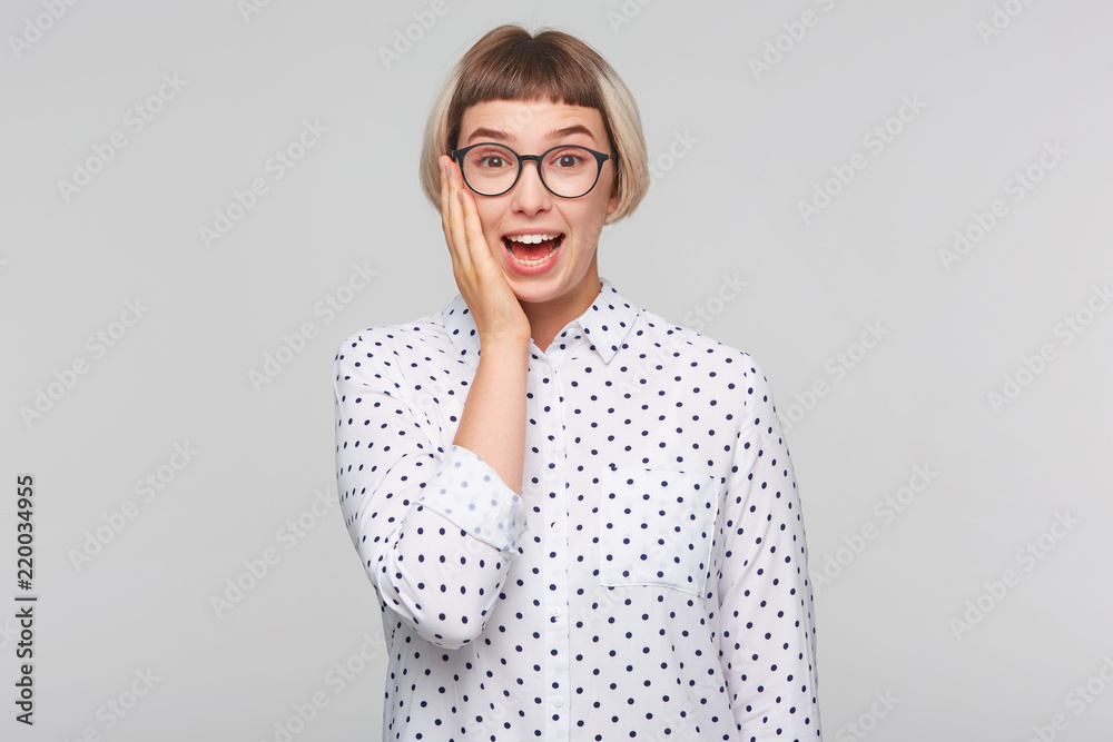 Closeup of cheerful amazed blonde young woman wears polka dot shirt and spectacles touching her cheek and looks feels surprised isolated over white background