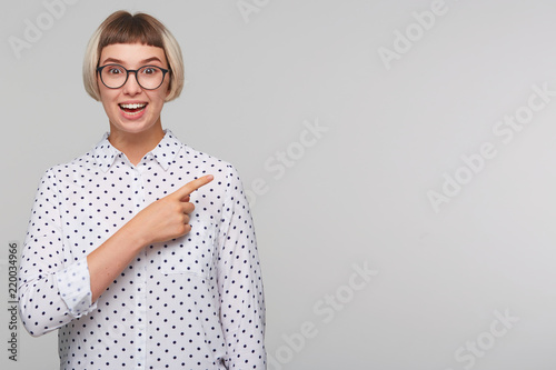 Closeup of happy beautiful blonde young woman wears polka dot shirt and spectacles feels excited and points to the side with finger isolated over white background