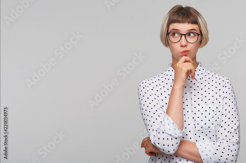 Portrait of pensive thoughtful blonde young woman wears polka dot shirt and glasses feels displeased and looks to the side isolated over white background photo