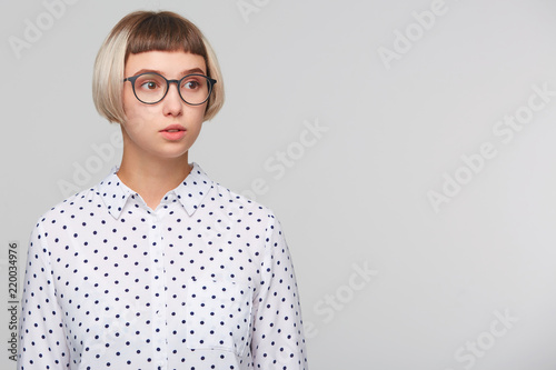 Portrait of pensive attractive blonde young woman wears polka dot shirt and glasses looks serious and looks to the side isolated over white background © timtimphoto