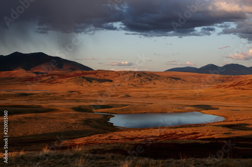 Colorful sunset above highland steppe shore of lake with dry yellow grass on the background of rocky mountains under storm sky