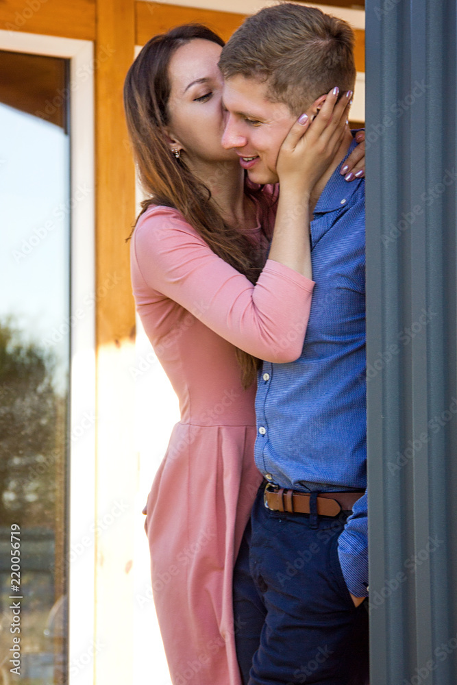 young happy pretty couple is standing and she kissing him to cheek on the wooden house background on the sunset picnic young happy pretty couple is standing and she kissing him to cheek on the wooden house background on the sunset picnic