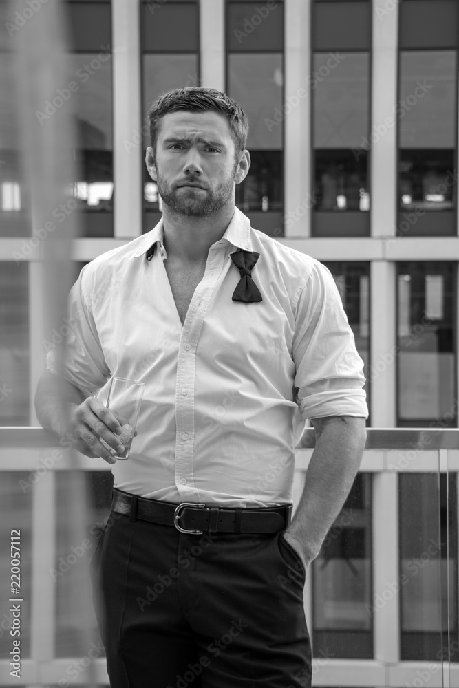 Handsome hunky man with unbuttoned shirt and loose bowtie stands on hotel balcony with sckyscraper backdrop Handsome hunky man with unbuttoned shirt and loose bowtie stands on hotel balcony with sckyscraper backdrop