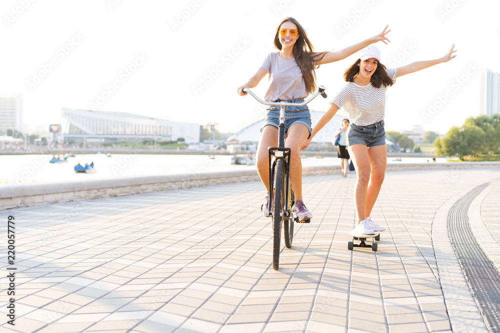Fototapeta premium Cute laughing young woman in jeans shorts on bike pulling girl holding on while using skateboard