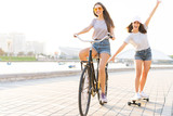Cute laughing young woman in jeans shorts on bike pulling girl holding on while using skateboard