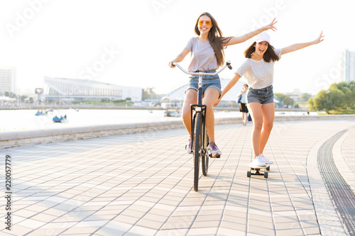 Cute laughing young woman in jeans shorts on bike pulling girl holding on while using skateboard
