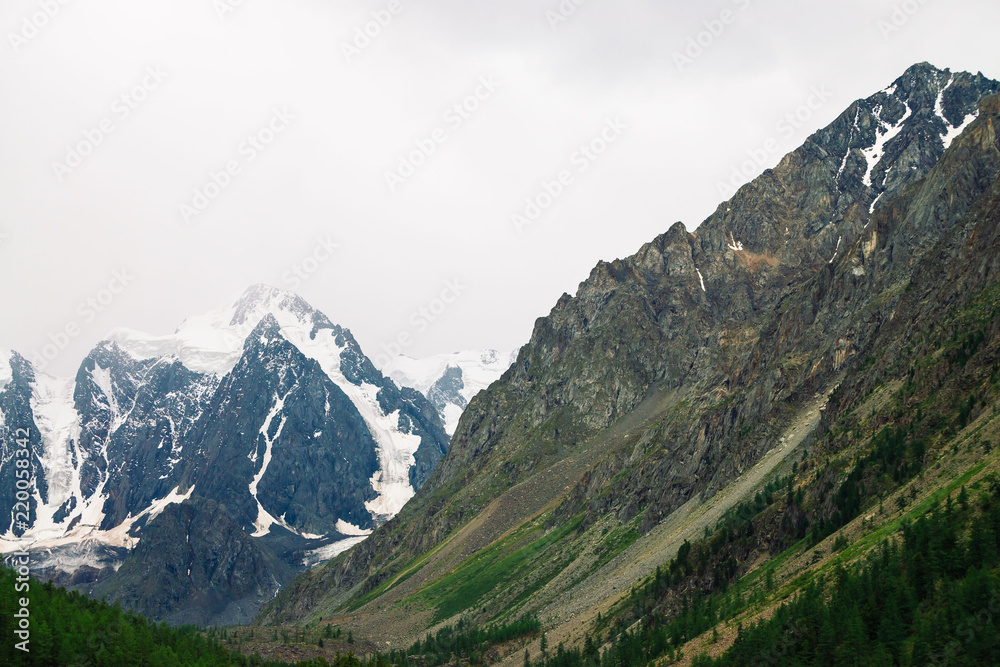 Snowy mountain top behind rocky mountain with forest under overcast sky. Rocky ridge in mist. Atmospheric minimalistic landscape of majestic nature.