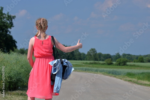 Nastolatka, blondynka, w letniej różowej sukience bez rękawów stoi tyłem na drodze i podniesionym kciukiem praej ręki daje znak, że podróżuje autostopem, w tle rozmyta zieleń, las