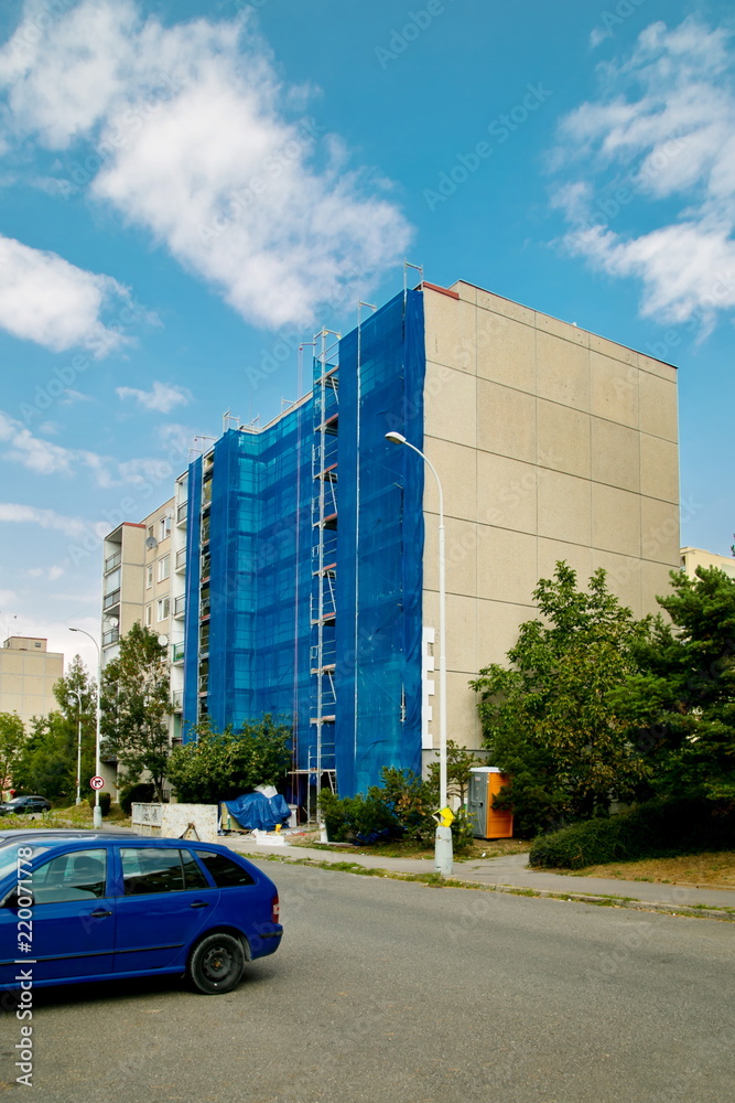 Thermal insulation, reconstruction work on a block of flats, blue canvas over scaffolding, street with cars, lamp, pavement, green tree, sunny blue sky, white clouds, vertical image