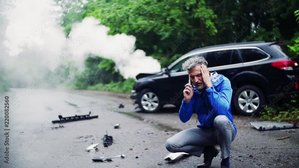 Mature man making a phone call after a car accident, smoke in the background.