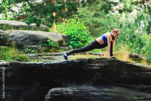 Blonde woman doing ashwa sanchalanasana outdoors on a rock in the forest. Yoga nature concept. photo