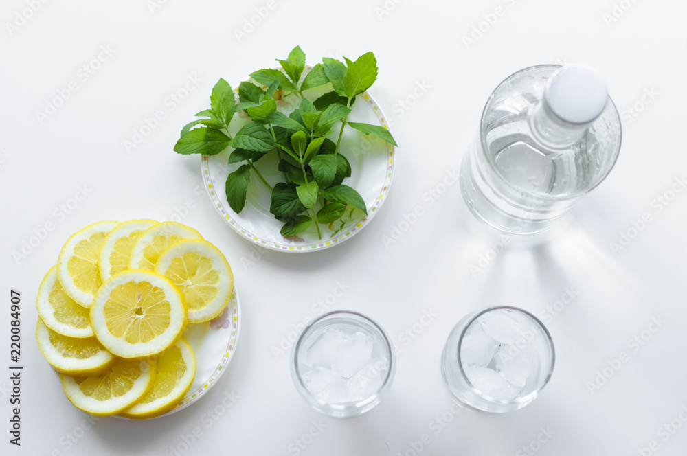 View from above on lemon slices on a plate, fresh mint leaves, bottle of water and two glasses with ice cubes. Lemonade set up on white background. View from above on lemon slices on a plate, fresh mint leaves, bottle of water and two glasses with ice cubes. Lemonade set up on white background.