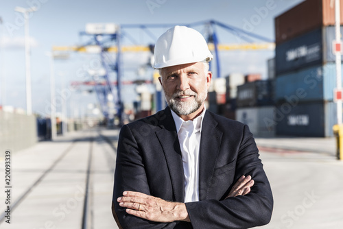 Businessman at cargo harbor, wearing safety helmet, arms crossed