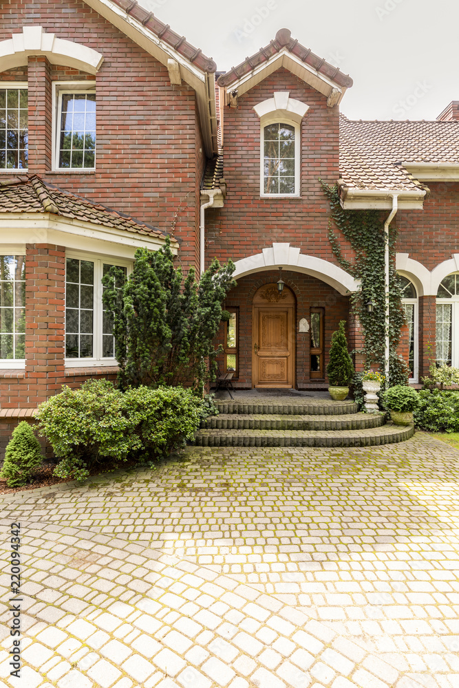 A welcoming entryway with ornamented wooden door, side windows and evergreens in a red brick stylish house. Empty cobbled path in a front.