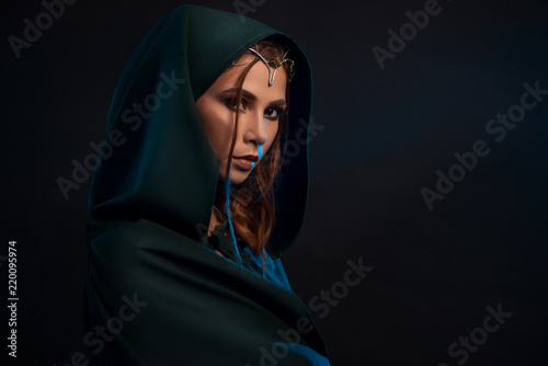 Fotografie Beatiful elf princess wearing dark green cape looking at camera from darkness