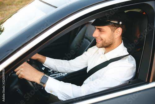 Portrait of happy european man taxi driver wearing uniform and cap, driving car fastening seat belt