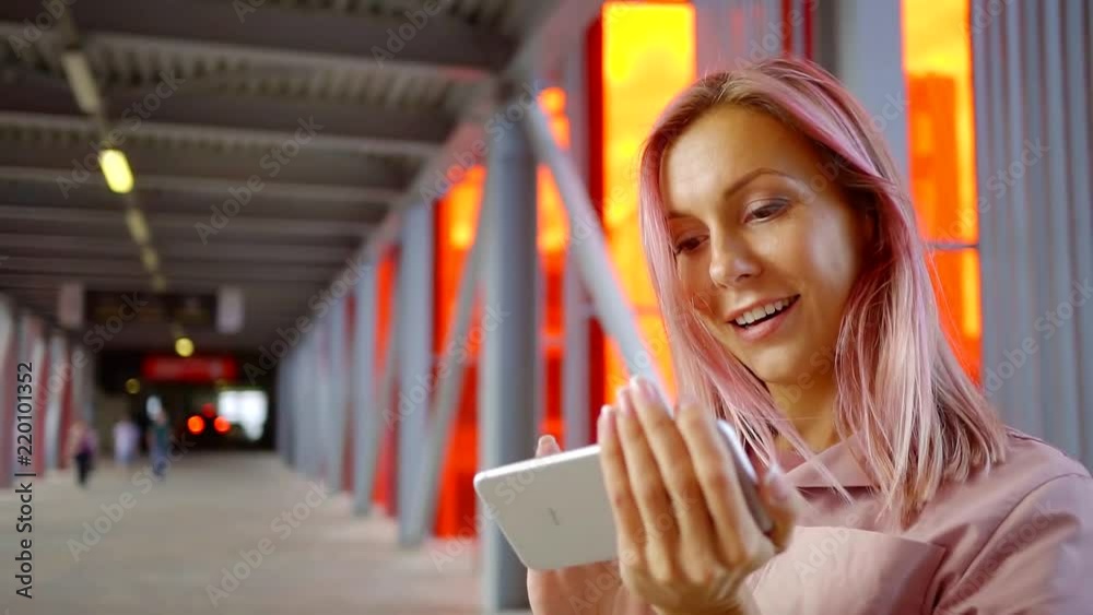 happy young girl with pink hair is standing in the Mall transition with a mobile phone in her hands and writing a text message