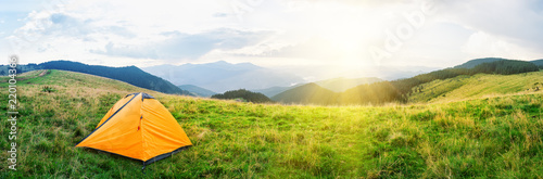 Orange tent on meadow with green grass in mountains under bright