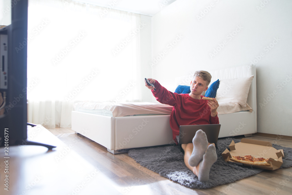 Young man sits on the floor in a cozy room with a laptop on his lap and pizza, switches the TV channels with the remote control and watches the TV. Work on a laptop with pizza and TV. Young man sits on the floor in a cozy room with a laptop on his lap and pizza, switches the TV channels with the remote control and watches the TV. Work on a laptop with pizza and TV.