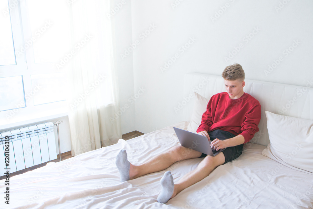 Young man sitting at home in a bright room on a bed with a laptop on his lap and working. Work at home on the Internet. Freelancer works on a laptop in his bed room Young man sitting at home in a bright room on a bed with a laptop on his lap and working. Work at home on the Internet. Freelancer works on a laptop in his bed room