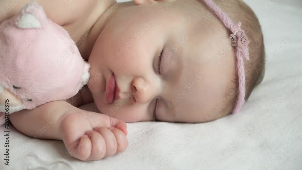 Little newborn baby girl with band sleeps with soft pink cat toy and pacifier, moves hand in dream. Concept of love and child care. Little newborn baby girl with band sleeps with soft pink cat toy and pacifier, moves hand in dream. Concept of love and child care.