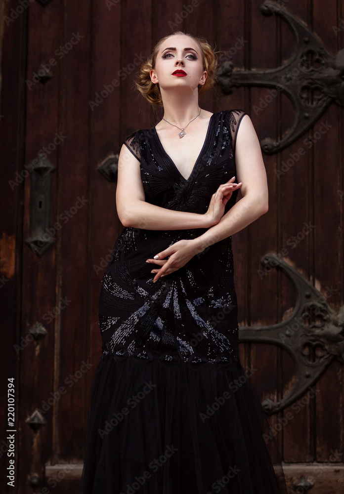 Portrait goth girl wearing black dress stands near red wood door .Beautiful witch outdoor, white skin red lips and blonde hair plaited .Halloween Vampire beautiful woman portrait Portrait goth girl wearing black dress stands near red wood door .Beautiful witch outdoor, white skin red lips and blonde hair plaited .Halloween Vampire beautiful woman portrait