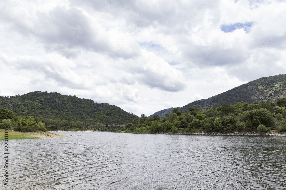view of a landscape of a lake surrounded by mountains and trees on a cloudy day
view of a landscape of a lake surrounded by mountains and trees on a cloudy day