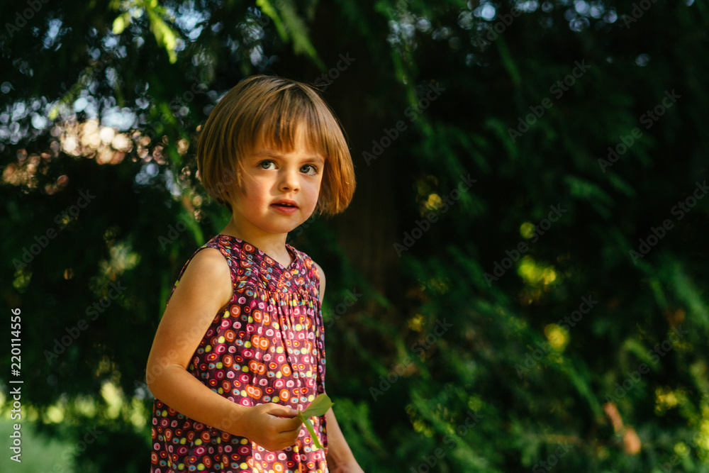 Cute baby playing outdoor in summer park alone. Three-year-old child in colorful dress on green background. Curious baby girl researcher touching a leaf with finger. Cute baby playing outdoor in summer park alone. Three-year-old child in colorful dress on green background. Curious baby girl researcher touching a leaf with finger.