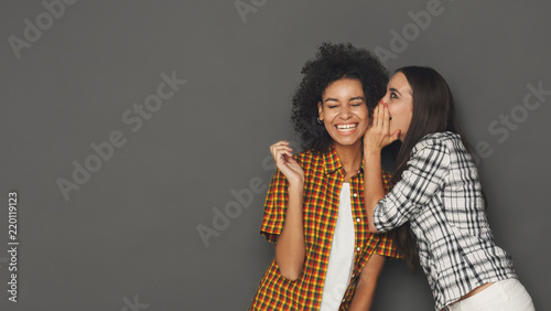 Young woman whispering to her friend against grey background photo