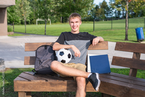 Happy teenager sitting on a bench with a soccer ball and his school supplies. photo