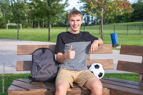 Happy teenager sitting on a bench with a soccer ball and his school supplies.