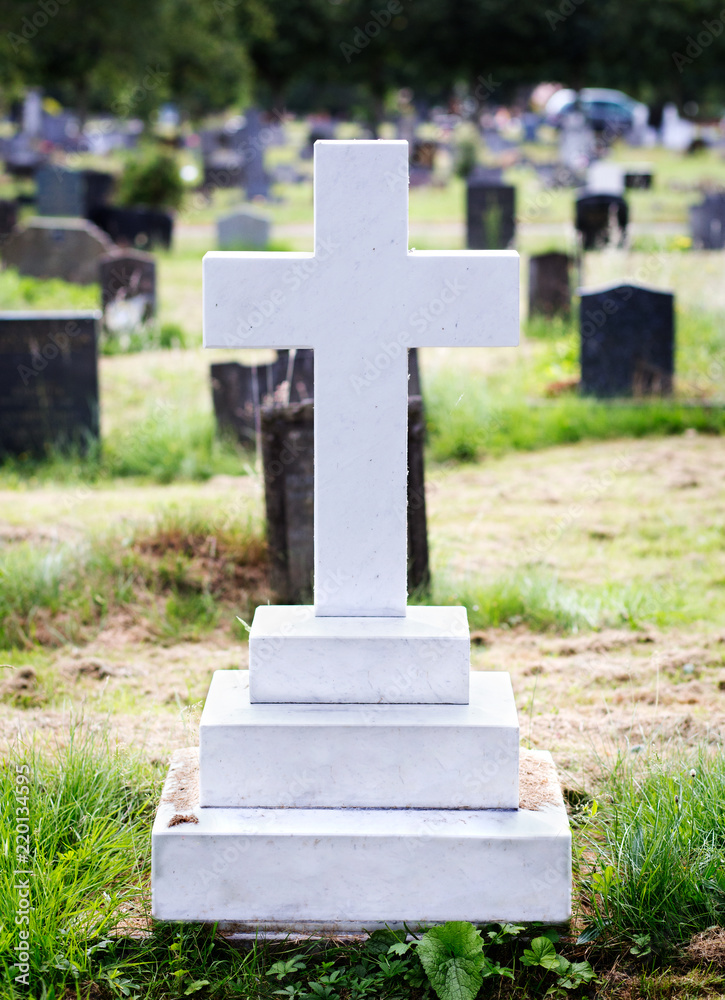 White marble gravestone (blank) in the shape of a cross. Trees and other graves in the background.
