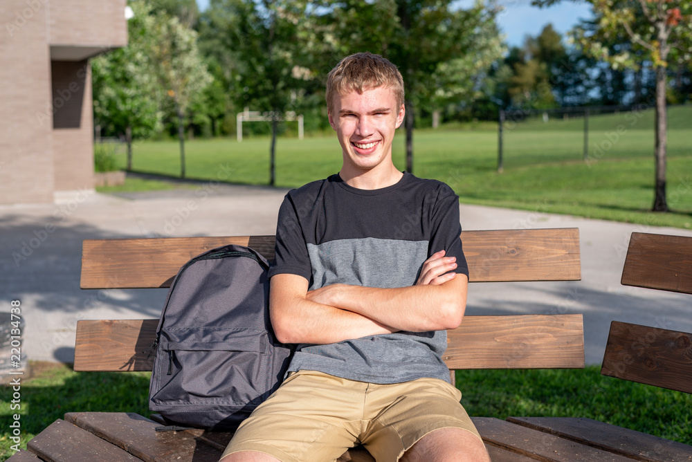 Fototapeta premium Happy teenager sitting on wooden bench that is next to a soccer/football field.