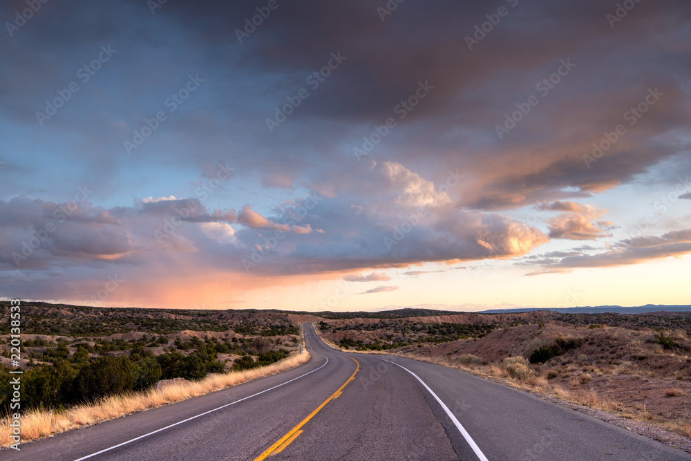 Naklejka premium Autostrada wijąca się w oddali przez krajobraz w pobliżu Santa Fe w Nowym Meksyku pod dramatycznym, kolorowym niebem o zachodzie słońca